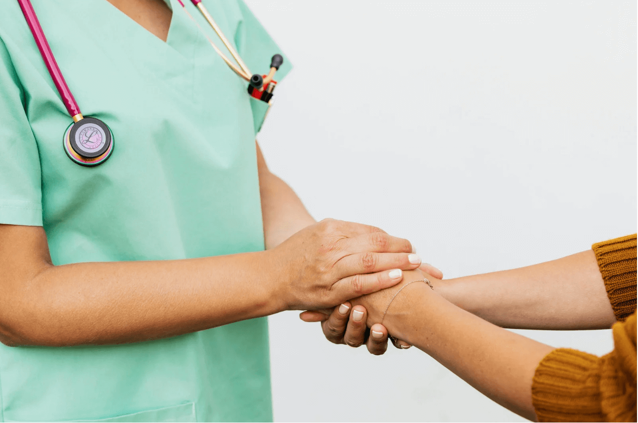 A nurse in green scrubs with a pink stethoscope holds a patient’s hand, showing care and support.