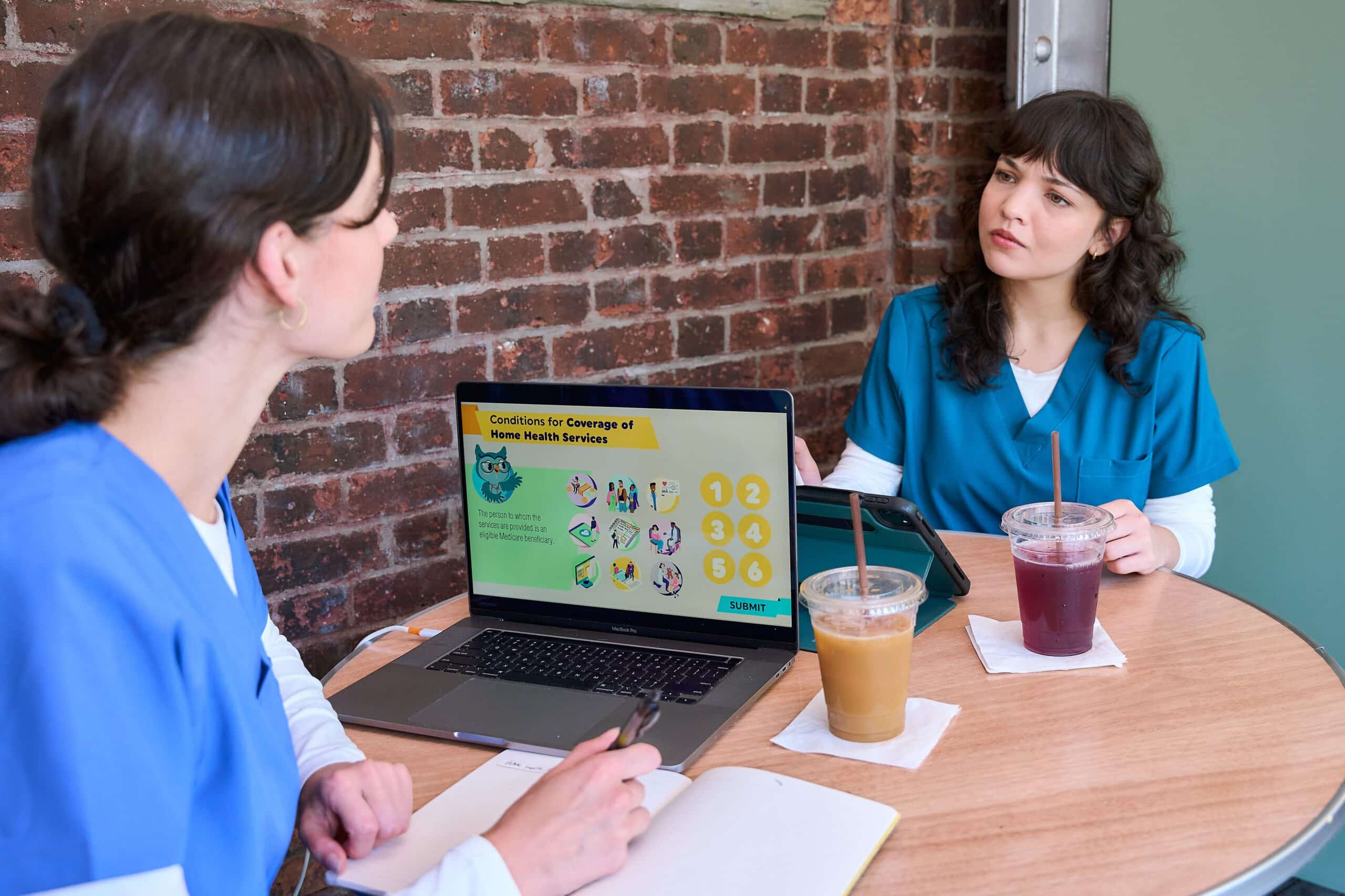 Two healthcare professionals in blue scrubs are sitting at a small round table with drinks, a laptop, and a notebook. The laptop screen displays a slide titled "Conditions for Coverage of Home Health Services" with colorful graphics and numbered icons. One person is taking notes while the other listens attentively.