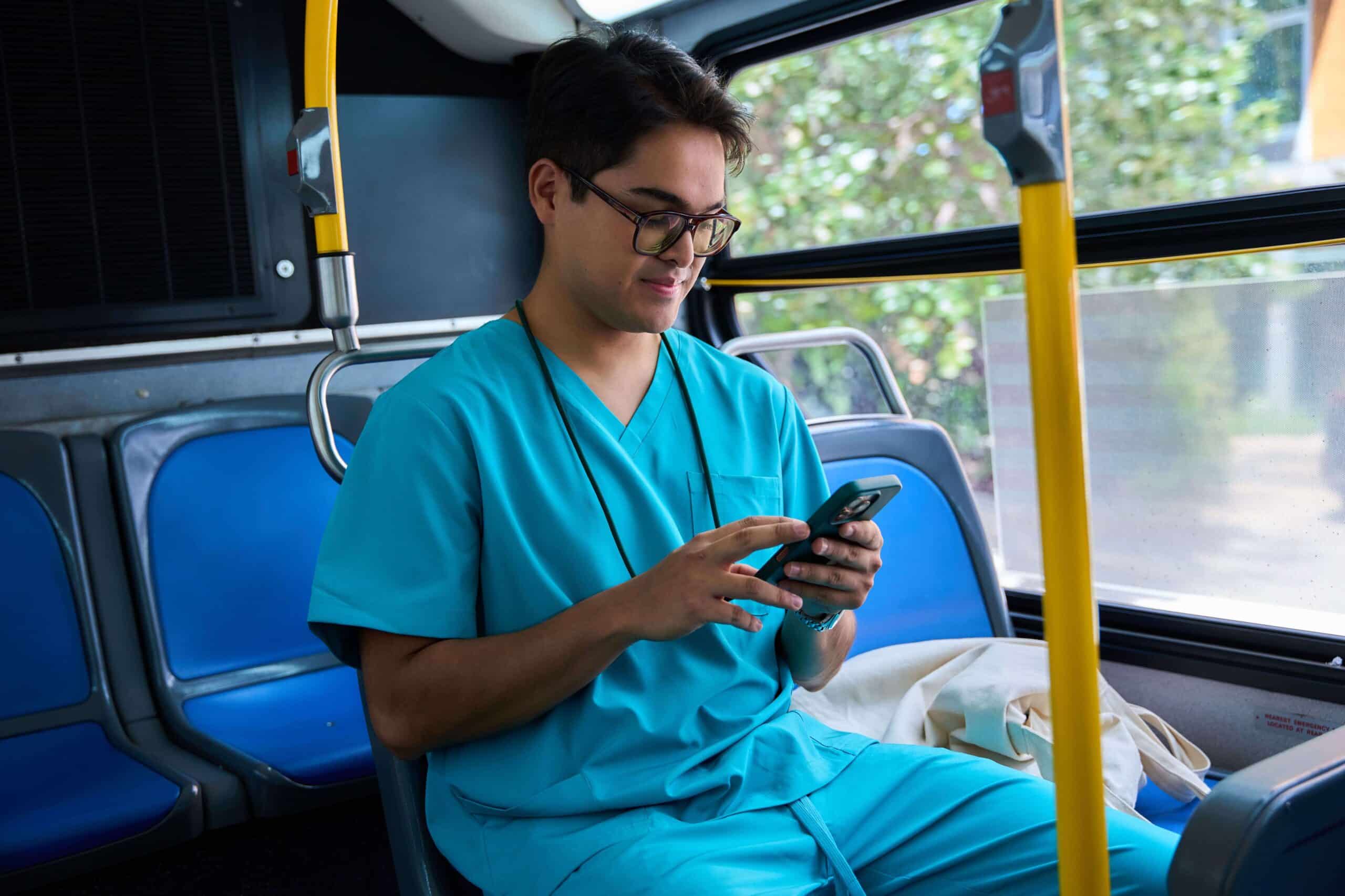A healthcare professional in teal scrubs sits on a bus, looking at their phone with a slight smile. They appear relaxed during the commute, with a tote bag placed beside them on the seat.