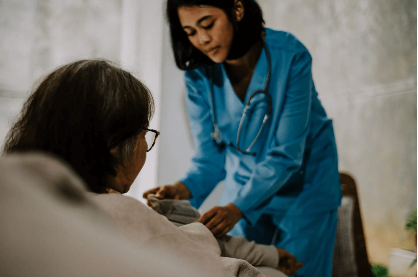A nurse in a blue uniform with a stethoscope assists an elderly woman in home care.
