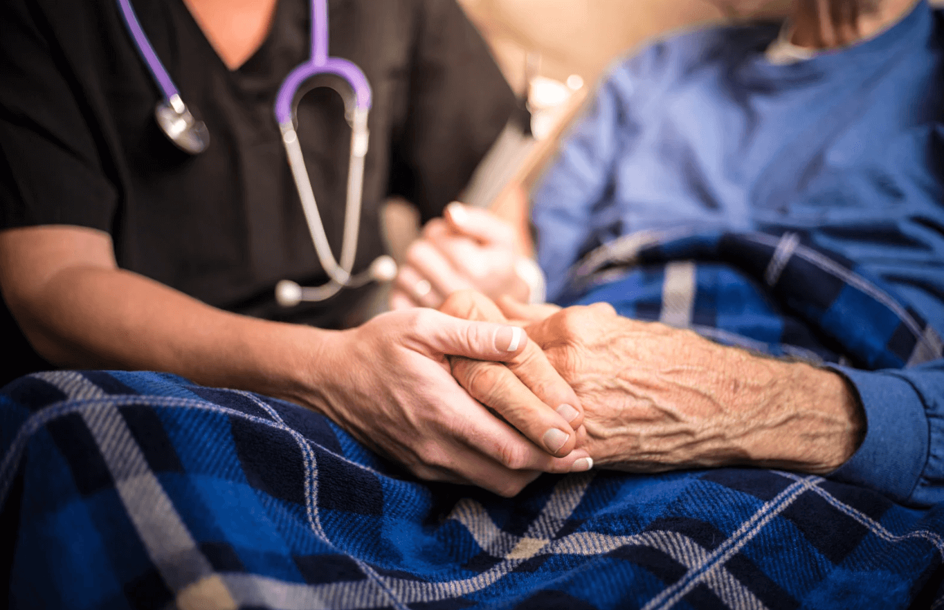 A healthcare worker with a stethoscope holds the hand of an elderly person covered with a blue blanket, showing compassion and care.