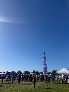 A crowd gathers on a sunny day at a Walk to End Alzheimer’s event, surrounded by tents and banners. A tall purple flag with the Alzheimer’s Association logo and the word “CARE” stands in the foreground.